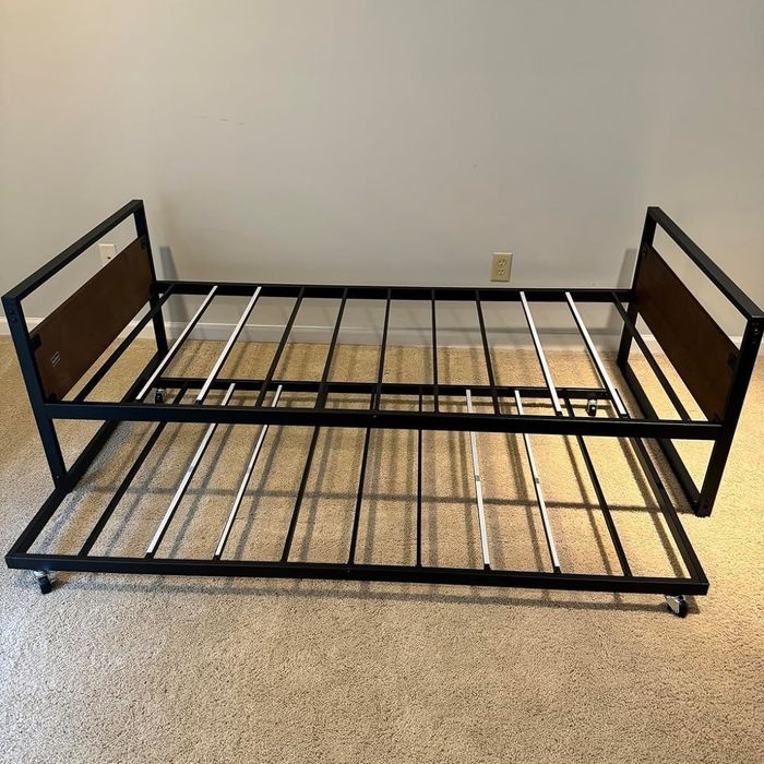 A metal twin trundle bed frame with wheels, displayed without a mattress, resting on a carpeted floor.