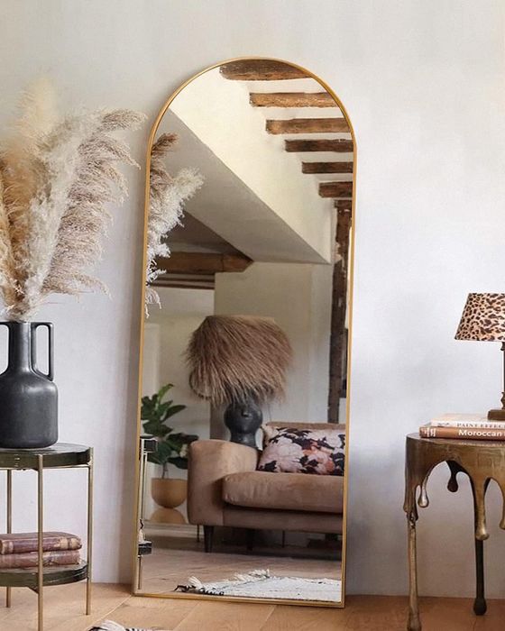 A full-length arched mirror in a cozy room with rustic furniture and plants, reflecting the warm ambiance of the interior. A lovely addition to home decor.
