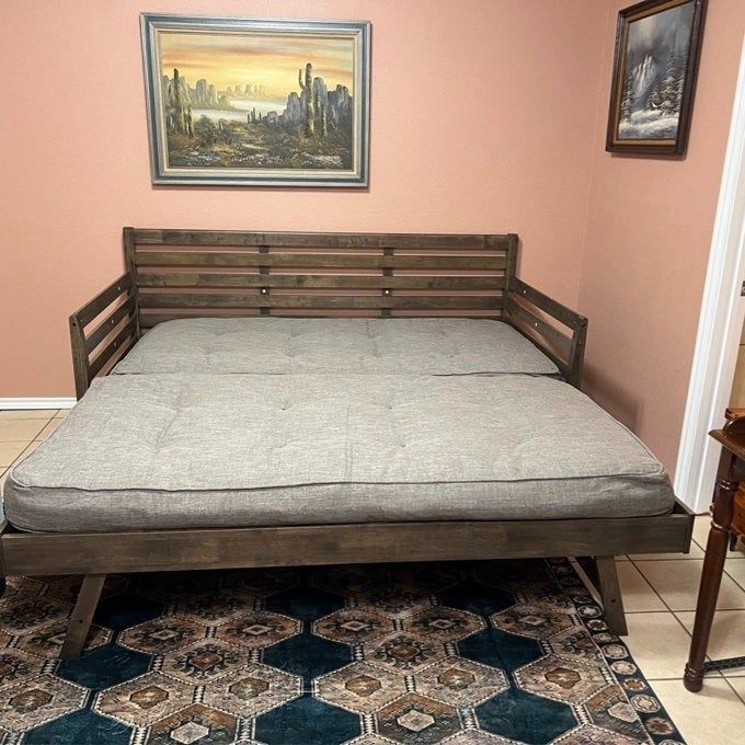 A wooden daybed with a gray mattress, positioned in a room decorated with southwestern and nature-themed paintings. A decorative geometric rug lies beneath it.