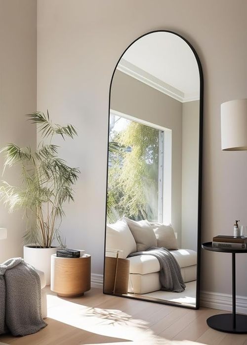 A tall arched floor mirror reflecting a cozy room with a charming nook, plants, and contemporary furniture, offering home decor inspiration.