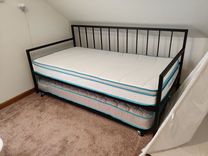 A twin-sized trundle bed with a sleek black metal frame, featuring a secondary mattress that slides underneath on a soft carpeted surface.