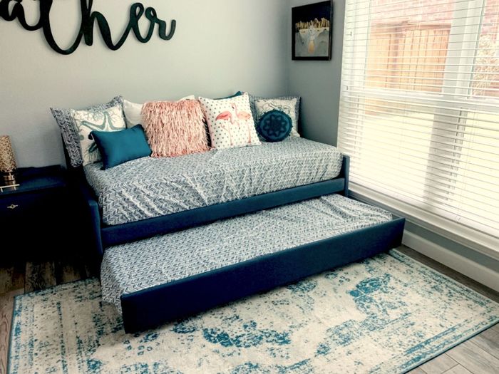 A daybed with a pull-out trundle in a snug room, accented by patterned pillows and a rug, with a large window nearby. The wall art above is partially visible.