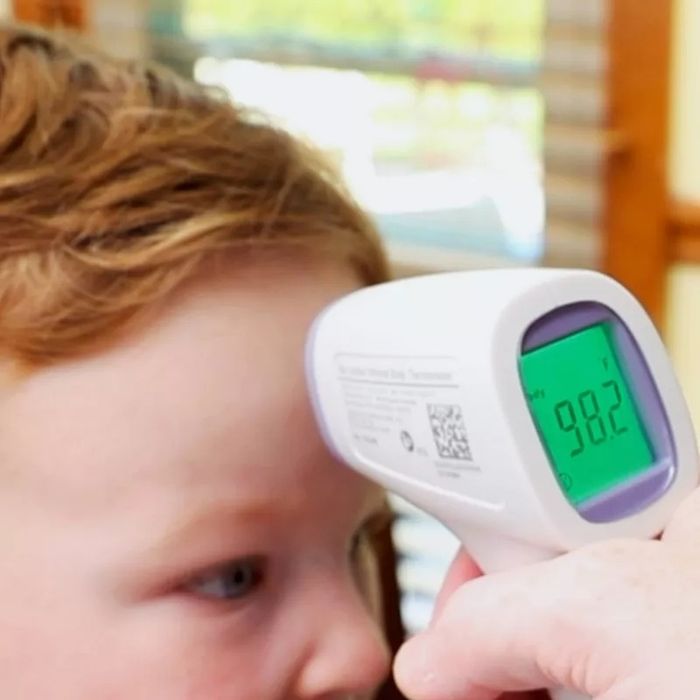 An individual uses a digital forehead thermometer, displaying a temperature of 98.2°F, aimed at a child’s forehead. The child is only partially visible in the image.