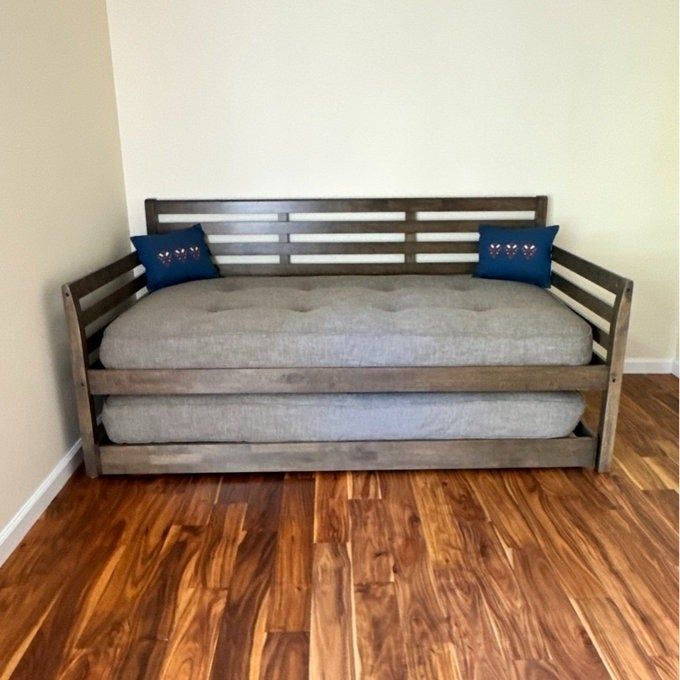 A wooden daybed with a sleek, slatted backrest and two blue pillows, positioned against a wall on a hardwood floor.