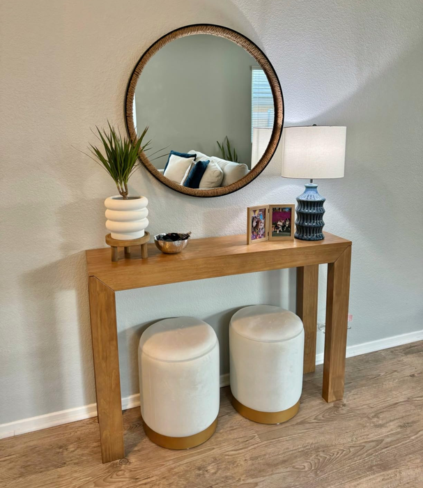 A round mirror above a console table, accompanied by decorative pieces and two ottomans below. Ideal for an entryway in any home.