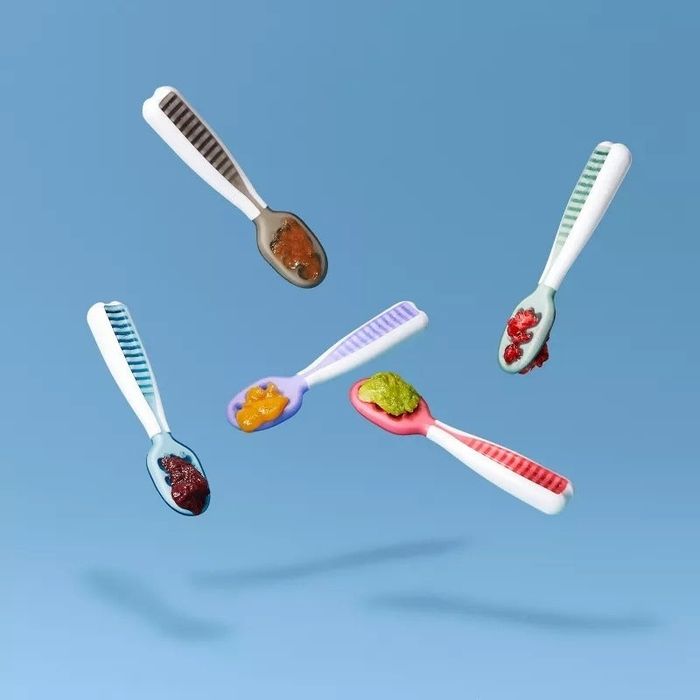 Five baby spoons with textured handles and various food samples, such as spreads and purees, are creatively suspended mid-air against a neutral backdrop.