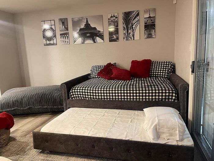 This cozy room features a daybed adorned with checked bedding and vibrant red cushions, complemented by a pull-out trundle and Paris-inspired wall art, all set against a stylish gray color scheme.