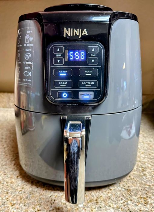 Reviewer's photo of a Ninja air fryer placed on a kitchen counter, showing 5:58 on the digital display, with functions like air fry, roast, and dehydrate clearly visible.