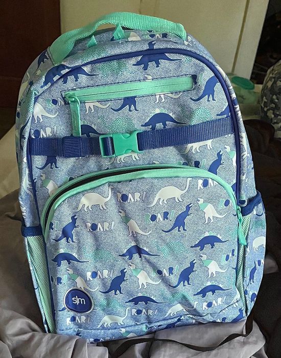 Reviewer's photo of a dinosaur-themed backpack featuring front and side pockets. The design includes dinosaur silhouettes and the text 'ROAR.'