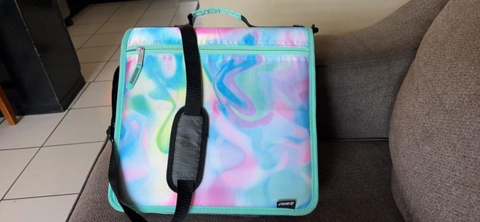 Reviewer's photo of the zipper binder in a pastel marble design, complete with a black shoulder strap, showcased on a gray sofa.