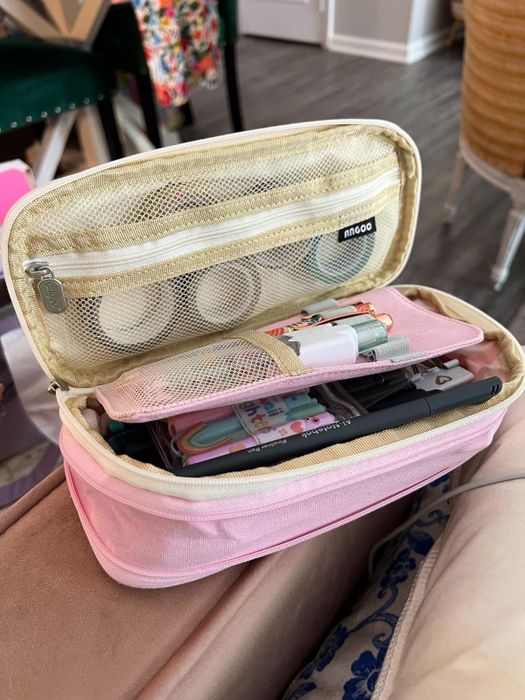 Reviewer's photo of an open pink pencil case with neatly arranged compartments, filled with stationery items like pens, markers, and rolls of washi tape, placed on a chair