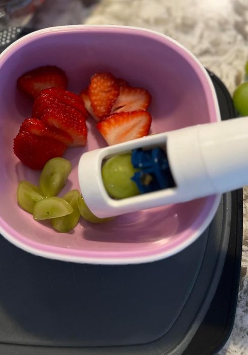 A reviewer’s photo showing sliced strawberries and green grapes in a pink bowl, with a white handheld grape slicer in action
