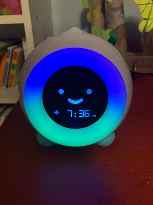 A reviewer's photo of the digital alarm clock featuring a cheerful face showing the time as 7:36 PM. In the background, a bookshelf filled with children's books is visible.