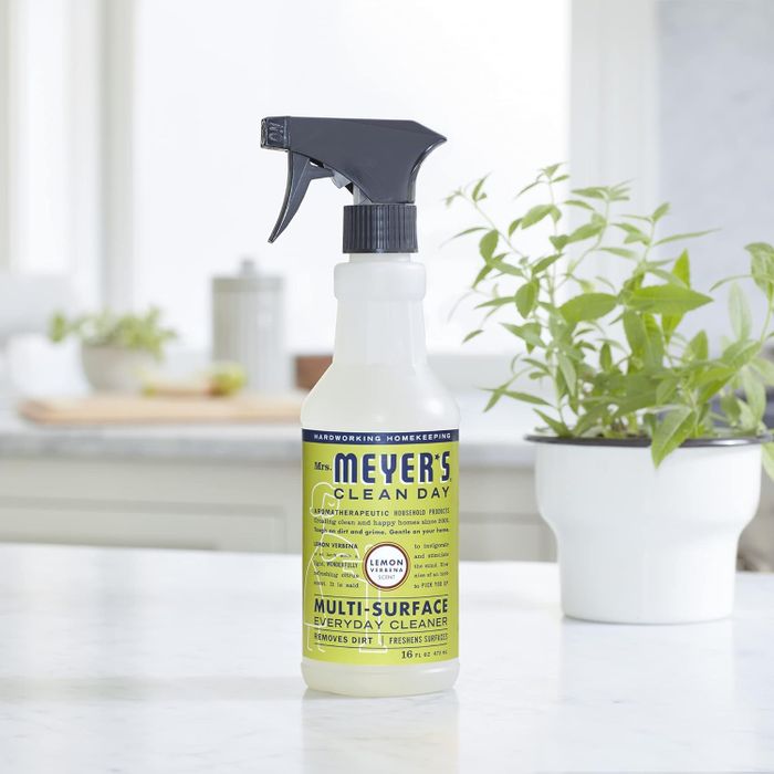 A bottle of Mrs. Meyer's Clean Day Multi-Surface Everyday Cleaner with lemon verbena scent, sitting on a kitchen counter next to a potted herb plant.