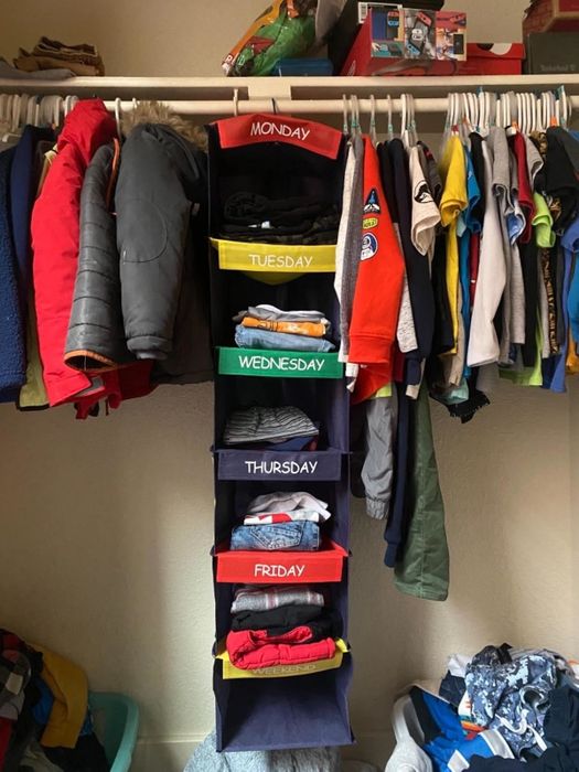 Reviewer's photo of a child's closet showcasing the hanging organizer labeled Monday through Friday plus Weekend, with various clothing items neatly placed in each section and additional garments hanging on the rod above.