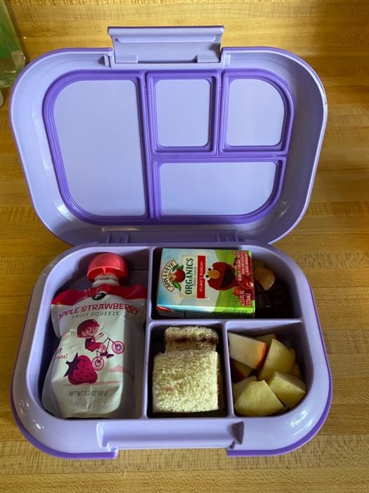 A reviewer’s photo of a purple Bentgo lunch box with four compartments, containing apple strawberry puree, an apple juice box, sandwich squares, apple slices, and gummies