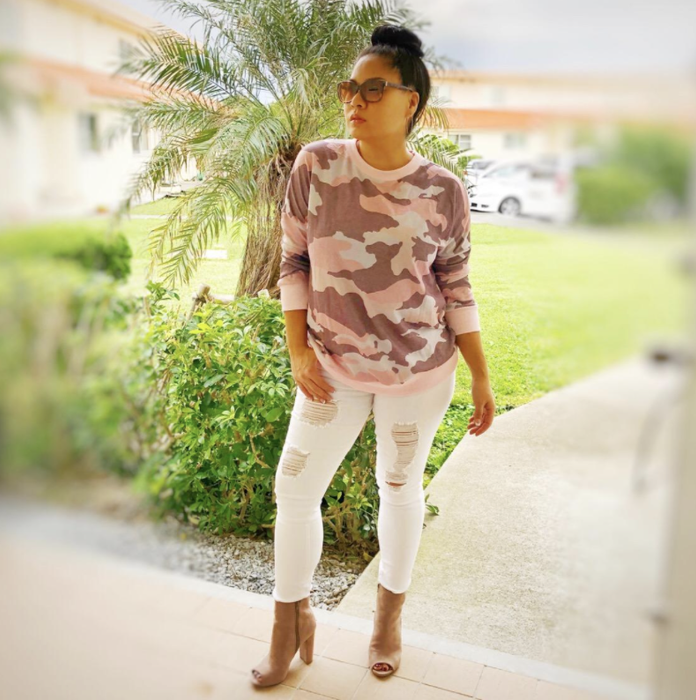A reviewer is seen wearing a pink camo print sweatshirt paired with white ripped jeans and ankle boots.