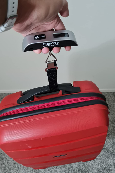 a reviewer using a digital luggage scale to weigh a red suitcase by attaching it to the handle and lifting it