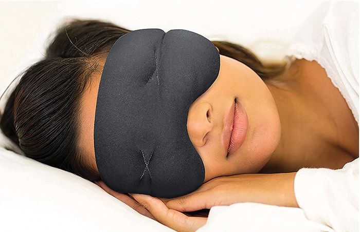 A model wearing a black sleep mask over their eyes, curled up in bed