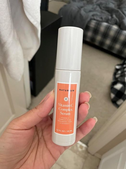A hand holds a 1 fl oz bottle of Naturium Vitamin C Complex Serum, containing a powerful combination of Vitamin C, hyaluronic acid, and Vitamin E to nourish and brighten skin.