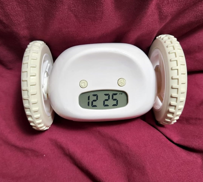 A white rolling alarm clock with a digital display showing 12:25, placed against a background of draped fabric.