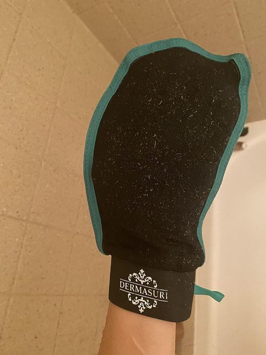A person is using the Dermasuri exfoliating mitt in a bathroom setting.