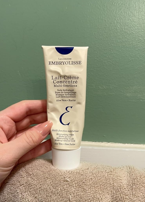 A hand holding a tube of Embryolisse Lait Crème Concentré Moisturizer against a green wall and beige fabric