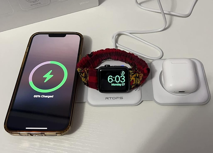 An iPhone charging at 68%, an Apple Watch showing the time 6:03 on its wristband, and AirPods resting on a wireless charging station.