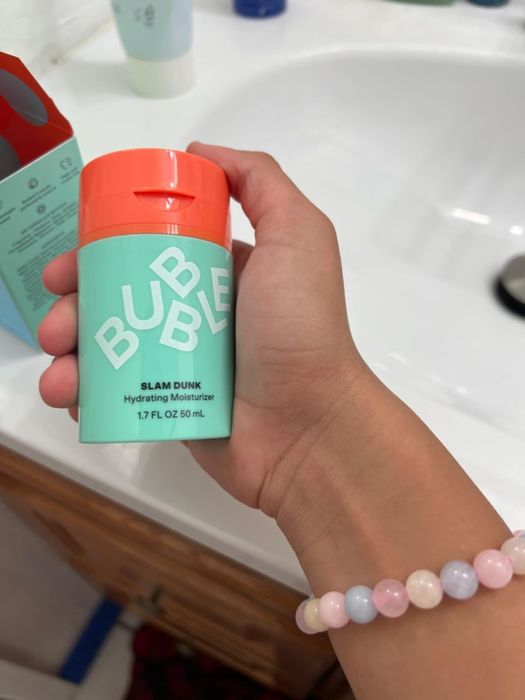 A hand holding the Billie Eilish-endorsed Bublé Slam Dunk Hydrating Moisturizer tube, 1.7 FL OZ, in a chic bathroom setting.