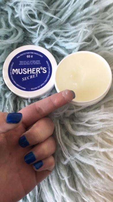 A finger applies Musher's Secret wax from an open container, with a blue-painted fingernail, set against a blurred background.