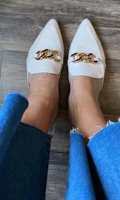 A close-up shot of someone wearing white pointed-toe loafers with a gold chain design on a wooden floor, paired with frayed blue jeans.