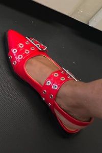 A person wearing blue jeans showcases a red flat shoe featuring metallic rings and buckles.