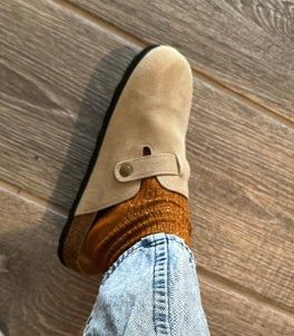 A close-up of a foot wearing a beige suede slip-on shoe with a buttoned strap, styled with textured socks and blue jeans.