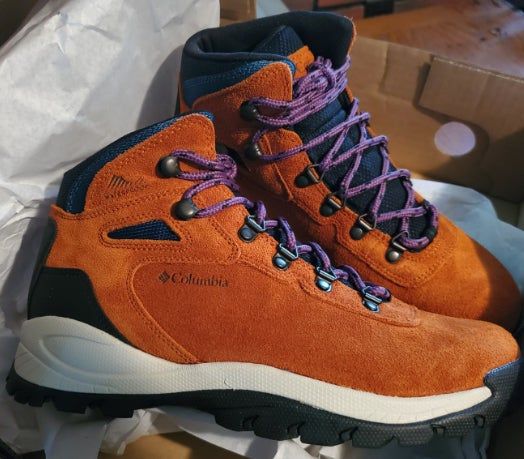 A pair of Columbia hiking boots, featuring orange suede and purple laces, displayed in a box, perfect for outdoor adventurers and hiking enthusiasts.