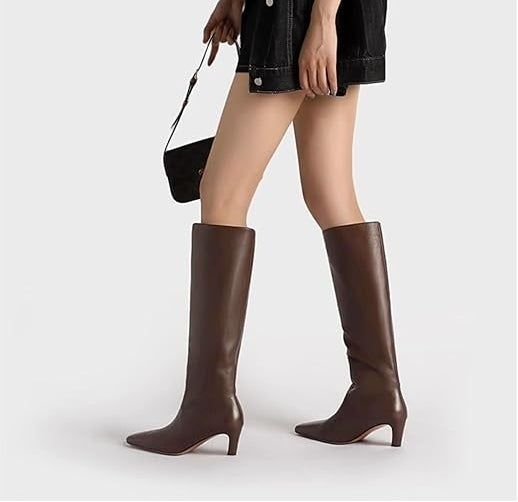 A person wearing knee-high brown boots, paired with an oversized black button-down shirt, holding a small black handbag.