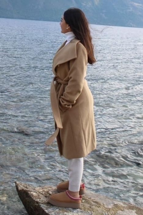 A reviewer standing by a lakeside, wearing a long camel-colored coat, white pants, and brown platform clogs, gazing thoughtfully into the distance.