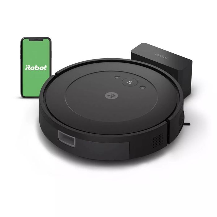 iRobot Roomba vacuum alongside its charging dock, with a smartphone displaying the iRobot app in the frame