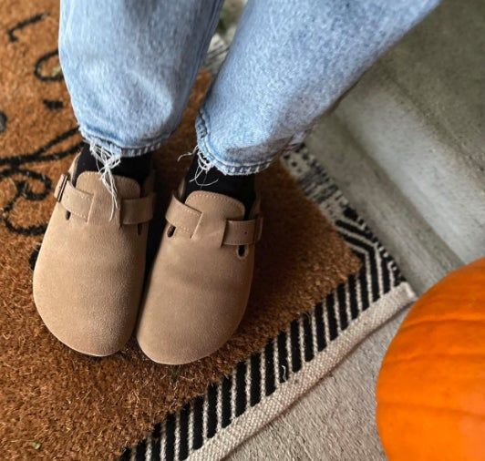 A close-up of a person's feet in light tan suede clogs, paired with frayed hem jeans. A pumpkin and a patterned rug are visible in the background.
