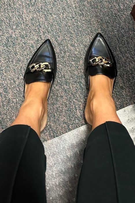 A person wearing black pointed-toe loafers with gold chain accents, paired with black pants, is seen in an indoor setting near a wooden desk.