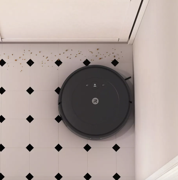 A robotic vacuum cleaner is sweeping crumbs along the edges of a tiled floor near a wall