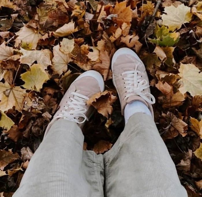 A person in corduroy pants and white sneakers, standing on a ground covered in fallen autumn leaves.