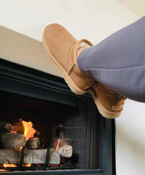 A person enjoys a cozy moment by the fireplace, sporting warm brown UGG boots for ultimate comfort.