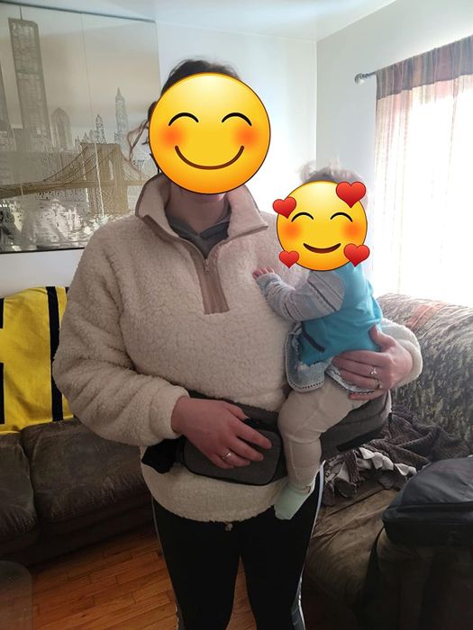 An adult in a comfy pullover carrying a joyful baby dressed in a blue outfit, with the Tushbaby attachment clearly visible, showcasing its practicality and comfort.