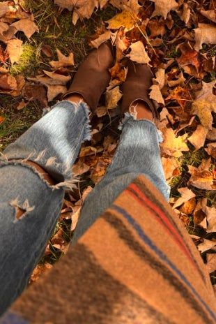 A person standing on a bed of fallen leaves, wearing ripped jeans and brown ankle boots.