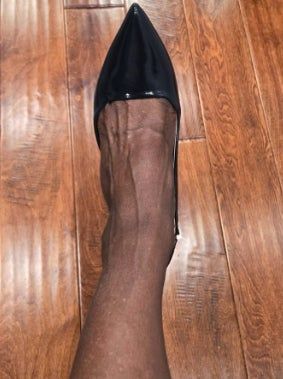 A close-up of a person’s leg wearing a shiny black pointed-toe high-heeled shoe on a wooden floor.