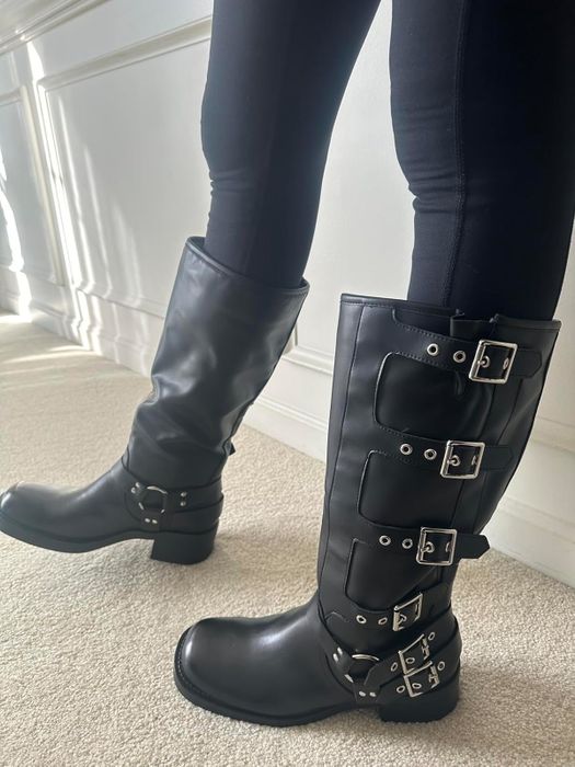 Close-up of a person wearing black knee-high leather boots with silver buckles, paired with black leggings.