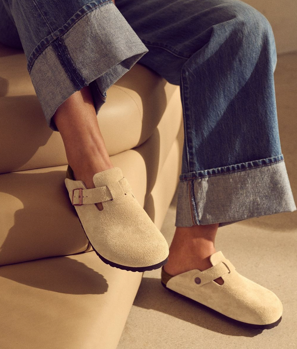 A person wearing wide-leg jeans and beige suede clogs with a buckle strap, sitting on a leather couch