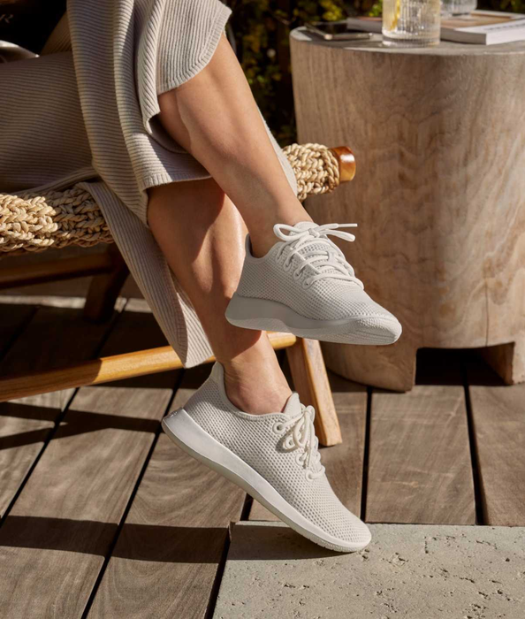 A person relaxing in a chair, wearing light-colored knit sneakers and a ribbed outfit, with a drink and a book resting on a nearby wooden table