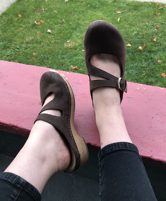 A writer showcasing brown backless clogs with buckled sides, featuring a wood and rubber sole