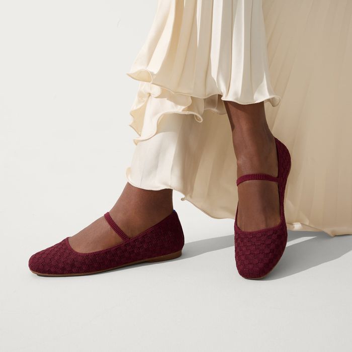 Close-up of a person wearing wine-colored knit ballet flats paired with a cream-colored, tiered, pleated dress. The shoes feature a strap across the top of the foot